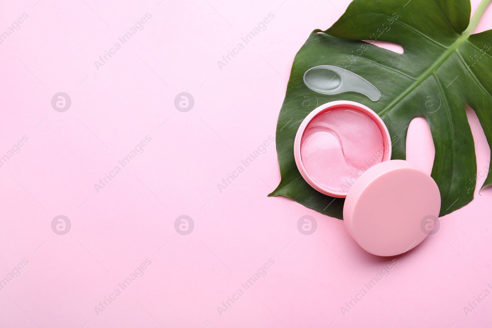 Photo of Under eye patches in jar with spatula and green leaf on light pink background, flat lay. Space for text