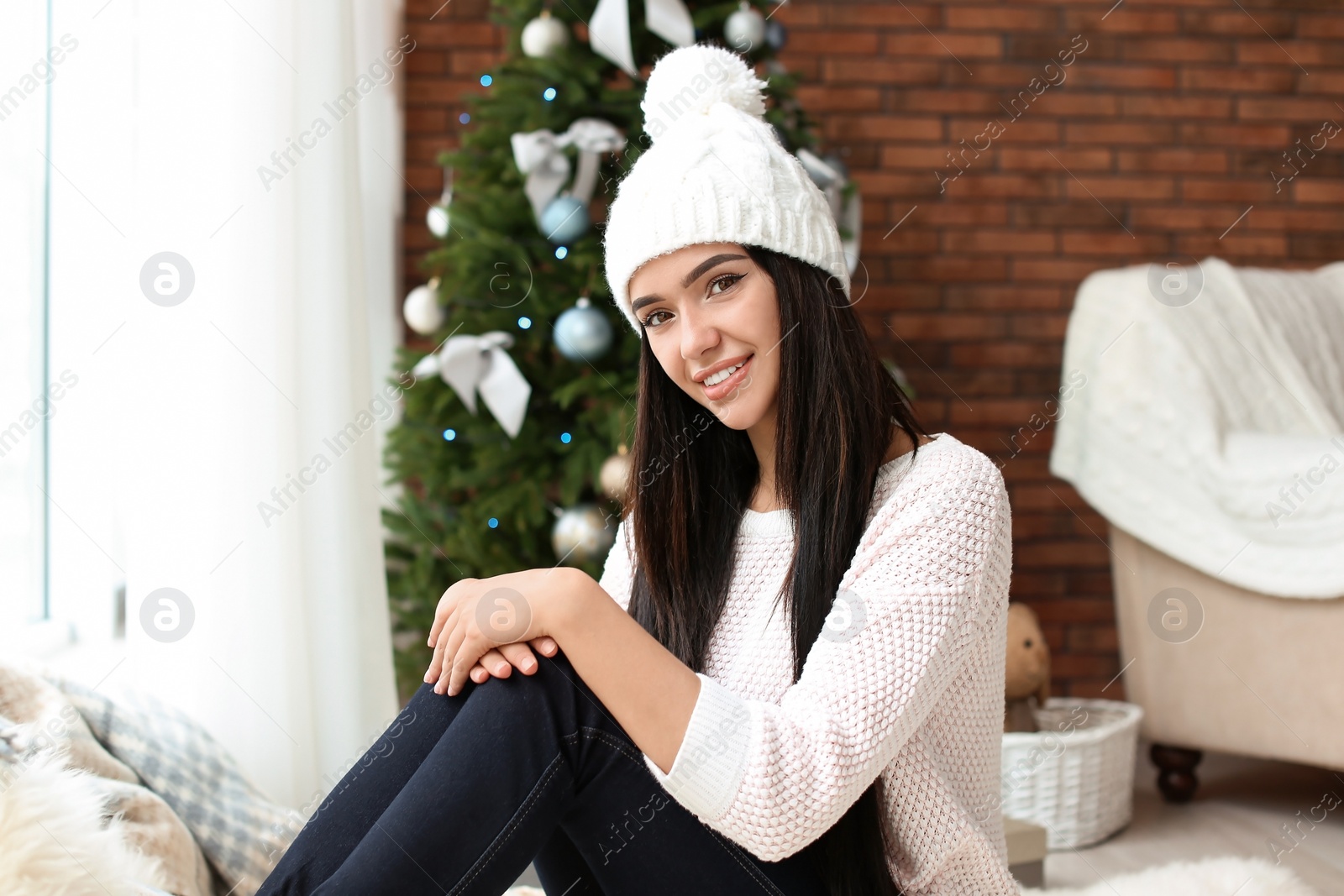 Photo of Beautiful young woman in hat near Christmas tree at home