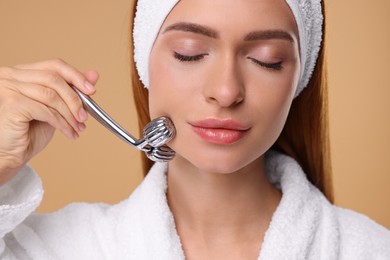 Photo of Young woman massaging her face with metal roller on pale orange background, closeup