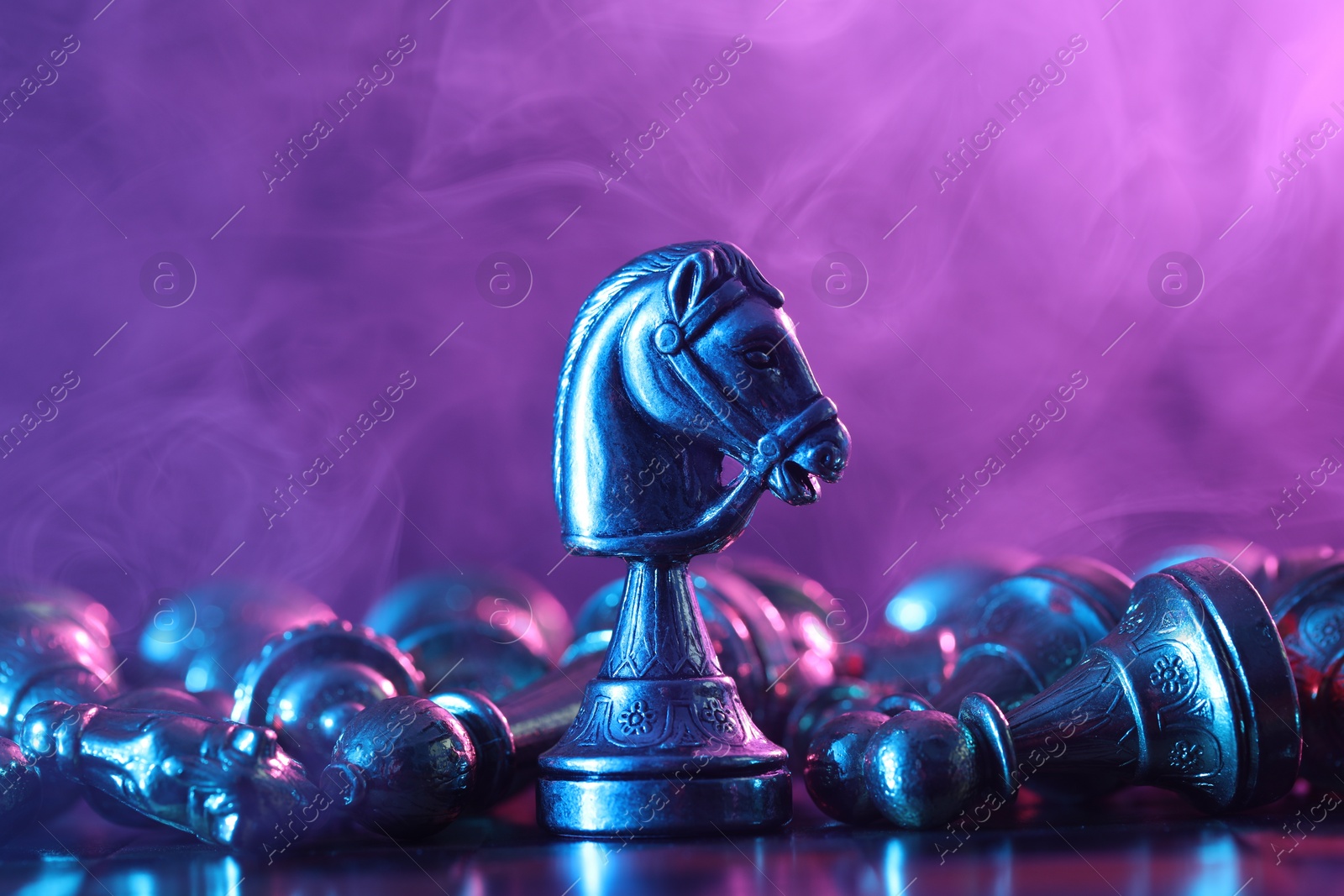 Photo of Metal knight among fallen chess pieces on checkerboard in color light, closeup