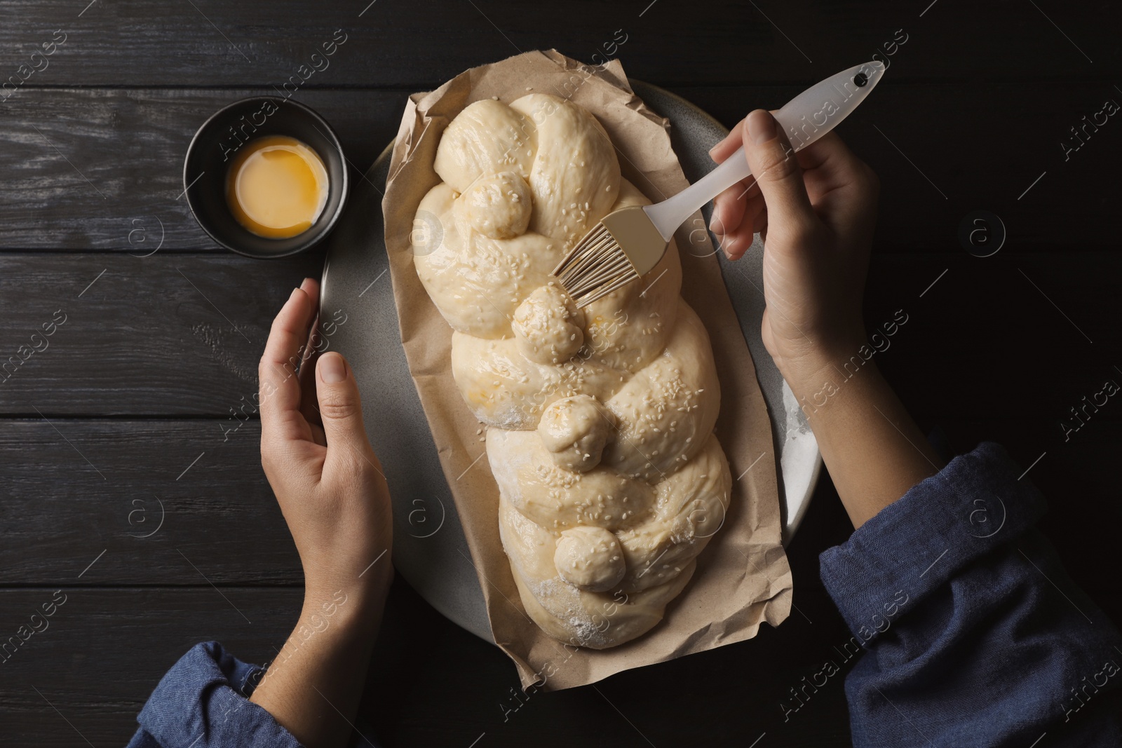 Photo of Woman spreading egg yolk onto raw braided bread at black wooden table, top view. Traditional Shabbat challah