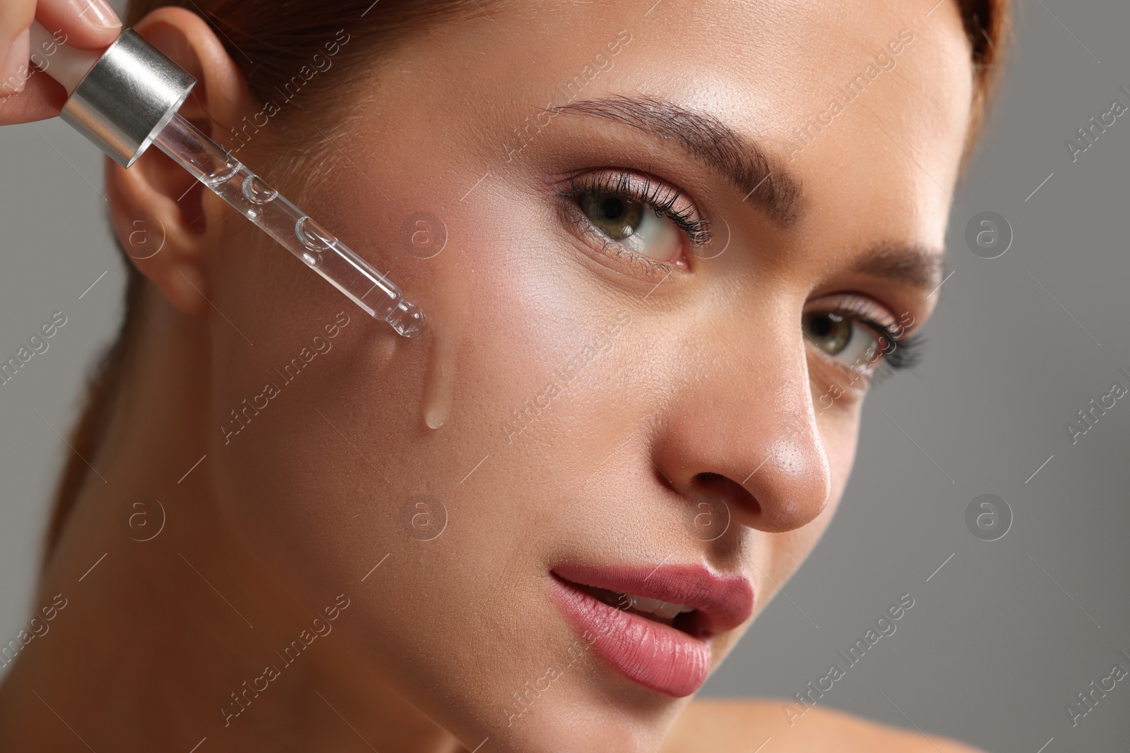 Photo of Beautiful young woman applying cosmetic serum onto her face on grey background, closeup