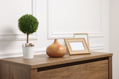 Green artificial plant in pot, frames and air humidifier on wooden chest of drawers near white wall