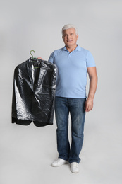 Senior man holding hanger with jacket in plastic bag on light grey background. Dry-cleaning service