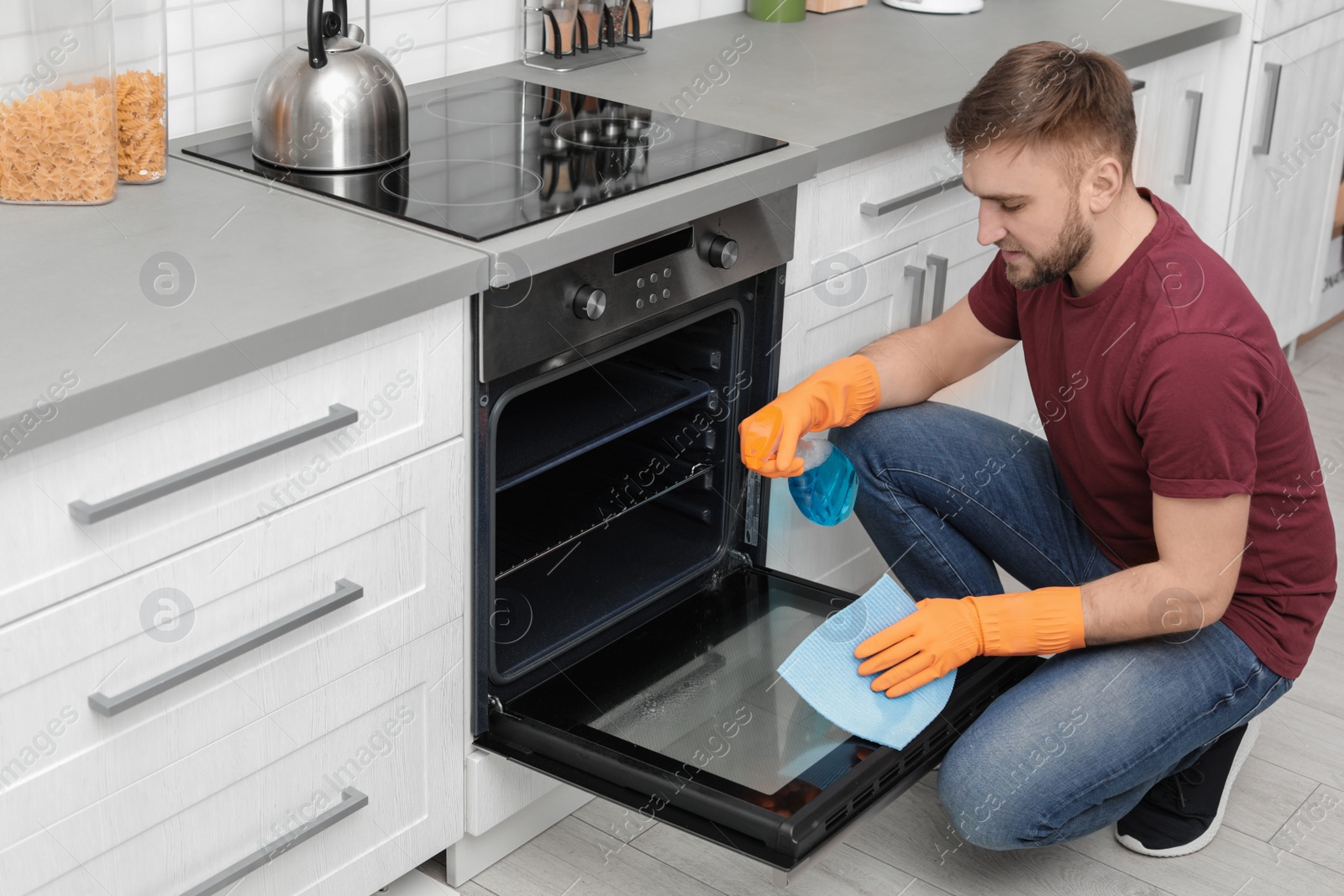 Photo of Young man cleaning oven with rag and detergent in kitchen