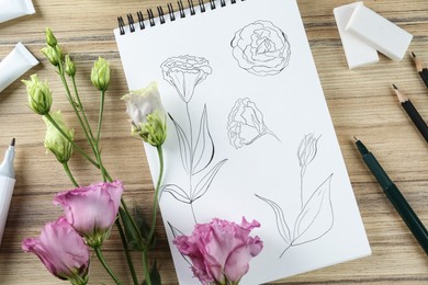 Photo of Sketches of flowers in notebook, eustomas and art supplies on wooden table, flat lay