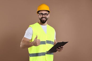 Engineer in hard hat holding clipboard and showing thumb up on brown background