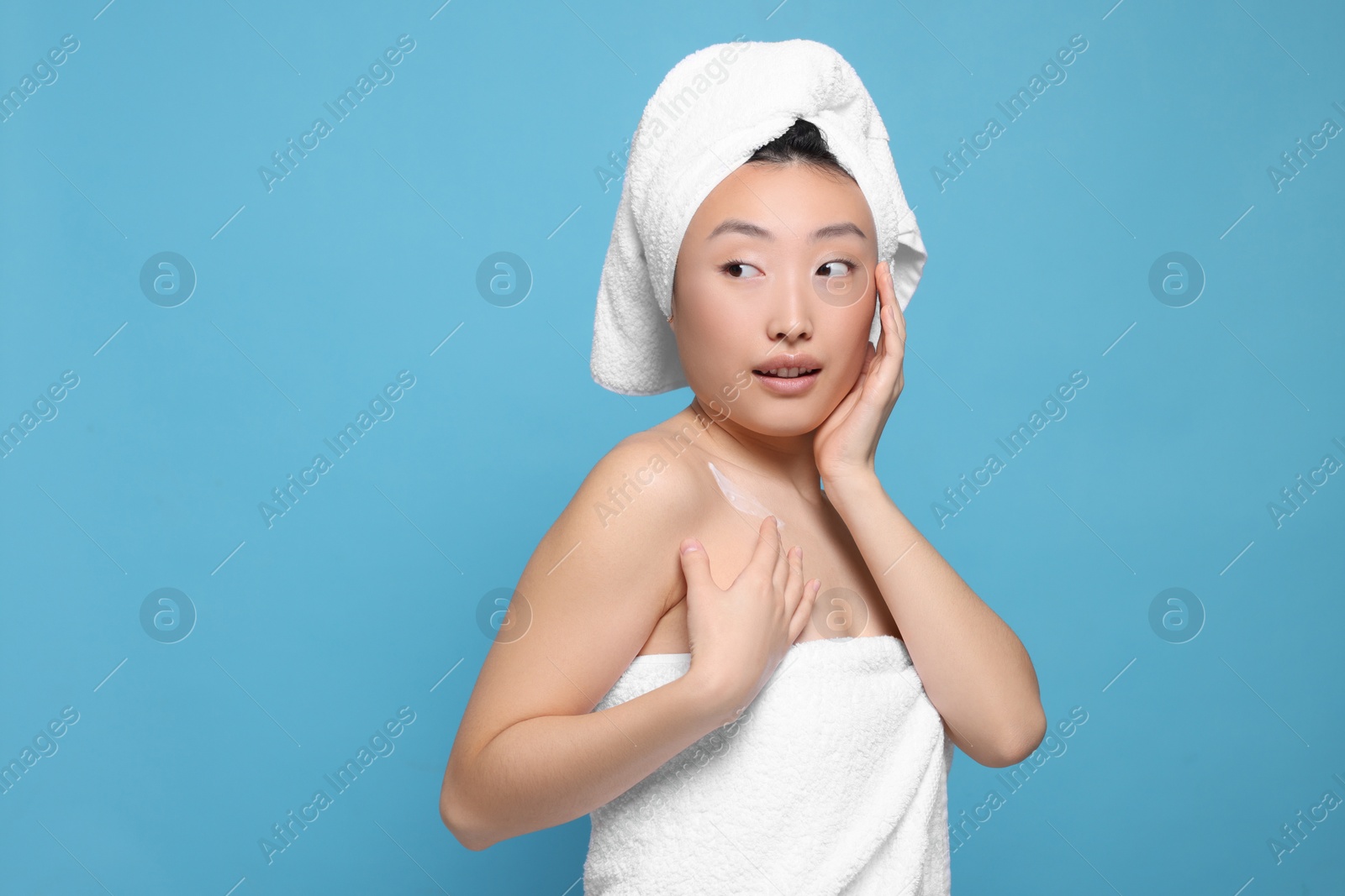 Photo of Beautiful young Asian woman applying body cream onto shoulder on light blue background, space for text