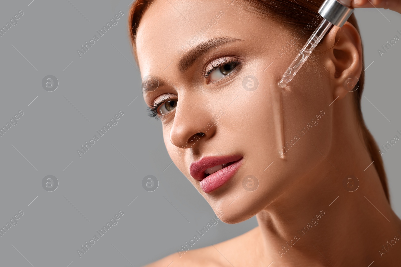 Photo of Beautiful young woman applying cosmetic serum onto her face on grey background, closeup. Space for text