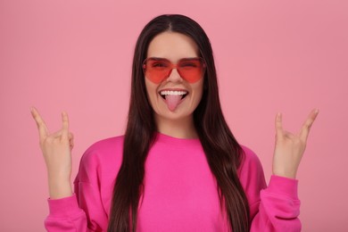 Photo of Happy young woman with heart shaped glasses showing her tongue and making rock gesture on pink background