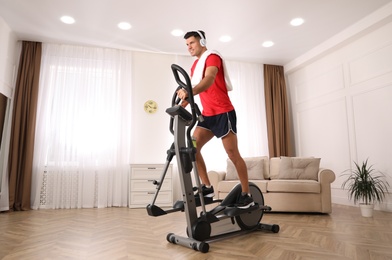 Photo of Man with headphones and towel using modern elliptical machine at home