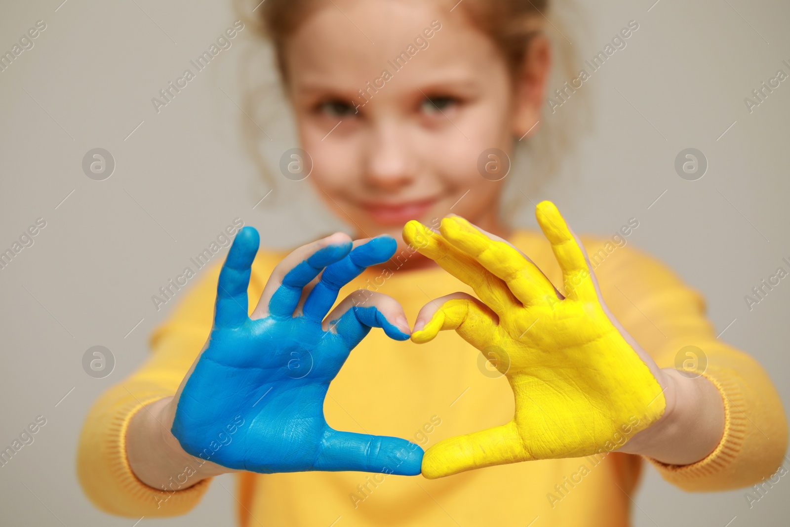 Photo of Little girl making heart with her hands painted in Ukrainian flag colors against light grey background, focus on palms. Love Ukraine concept
