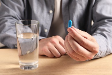 Man holding pill near glass of water at wooden table, closeup
