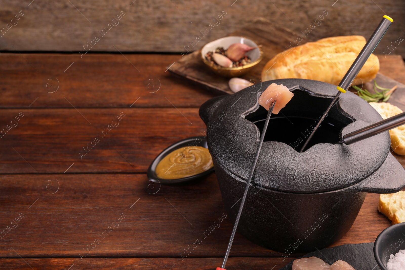 Photo of Fondue pot, forks, pieces of raw meat, bread and spices on wooden table, space for text