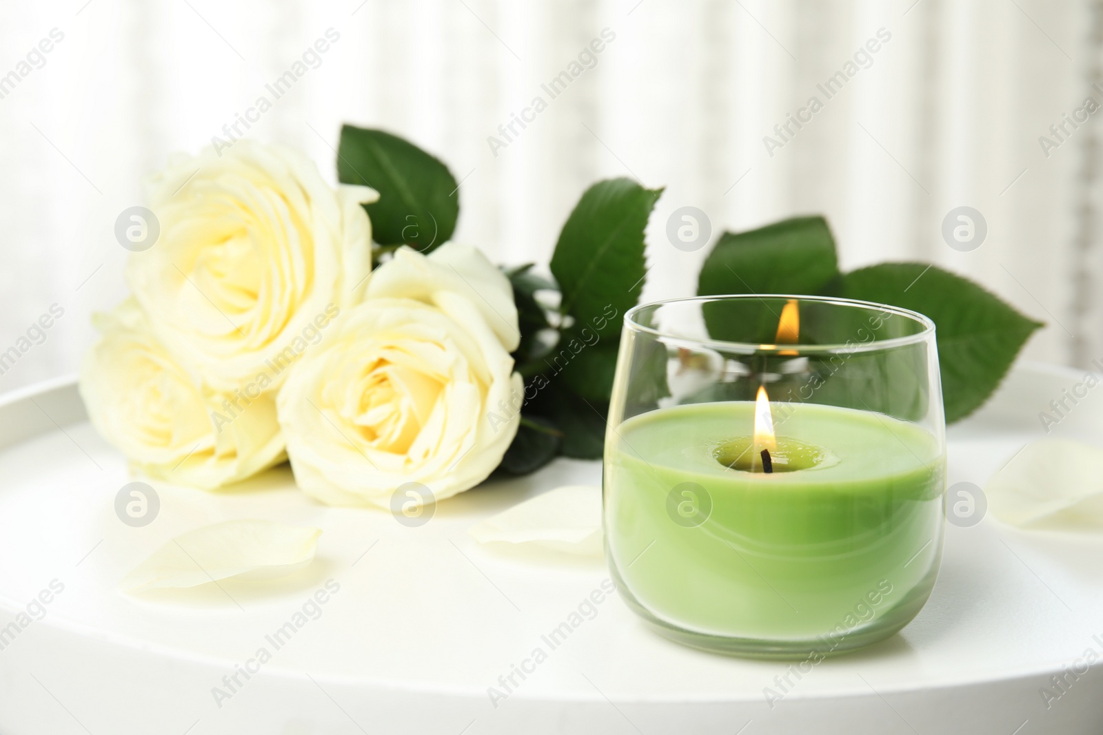 Photo of Burning candle in glass holder and roses on white table, space for text