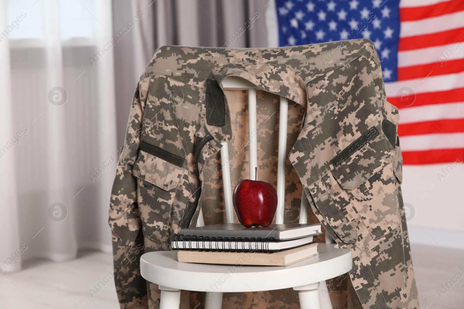 Photo of Chair with soldier uniform, notebooks and apple near flag of United States indoors. Military education