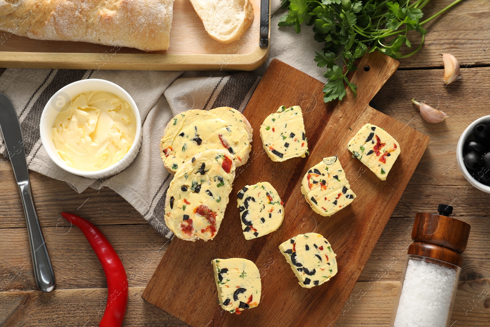 Photo of Tasty butter with olives, chili pepper, parsley and bread on wooden table, flat lay