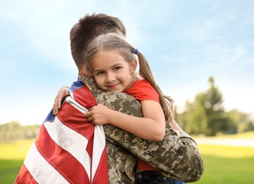 Father in military uniform with American flag hugging his daughter at sunny park