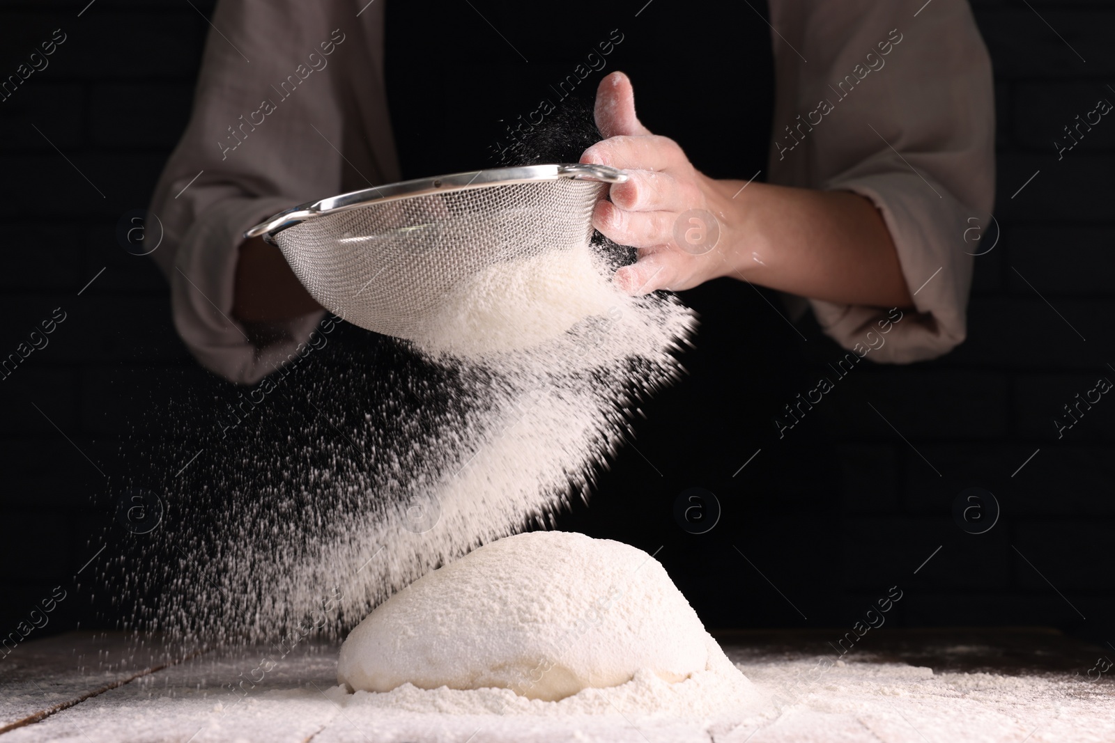 Photo of Woman sprinkling flour over dough at wooden table on dark background, closeup