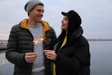Photo of Couple in warm clothes holding burning sparklers near river