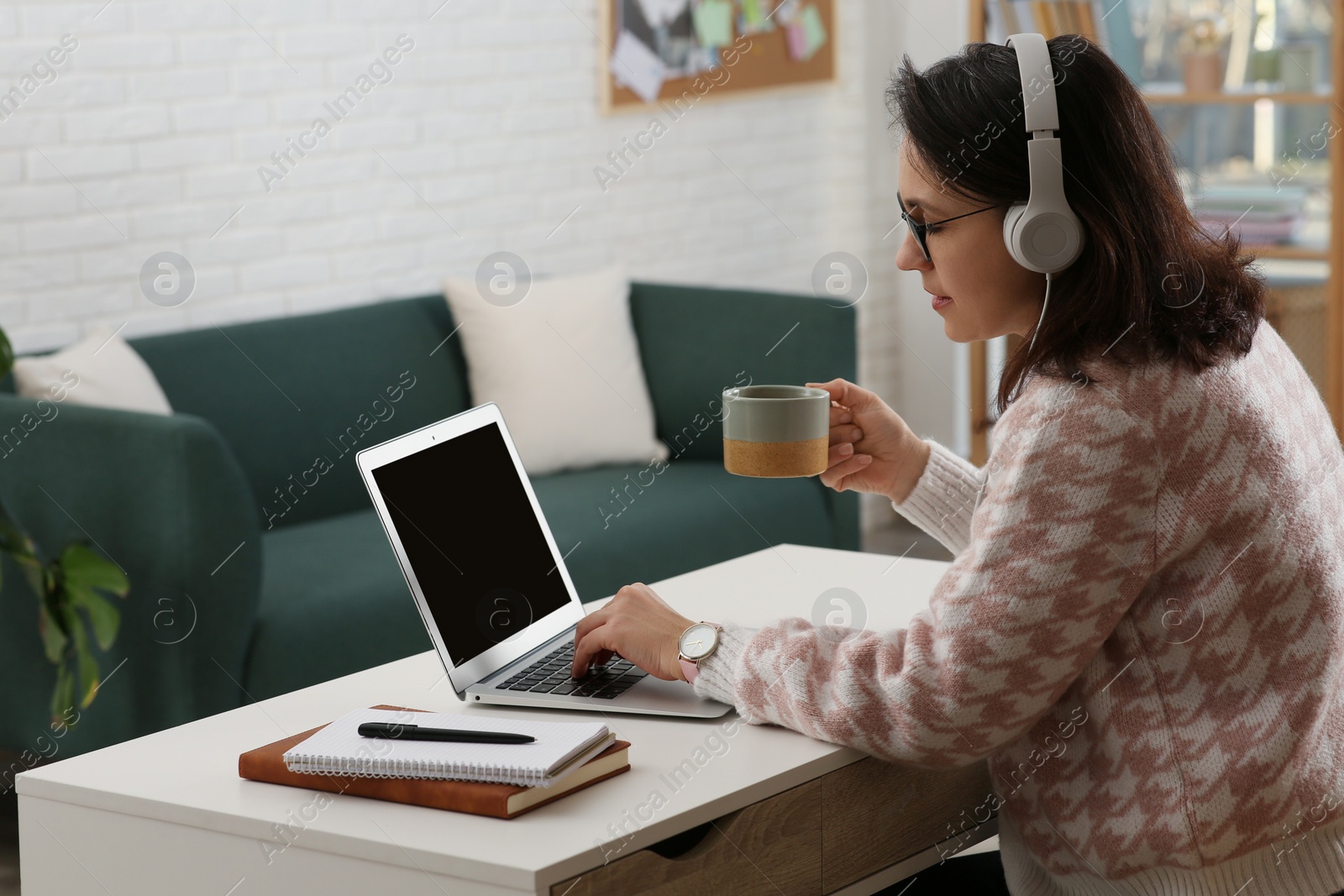 Photo of Woman with modern laptop and headphones drinking tea while learning at table indoors