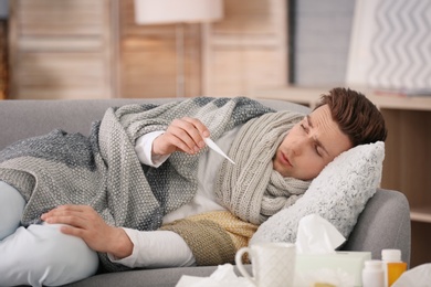 Photo of Sick young man with thermometer suffering from cold on sofa at home