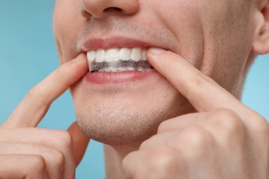Photo of Young man applying whitening strip on his teeth against light blue background, closeup