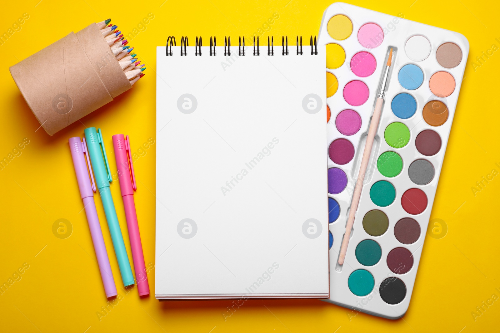 Photo of Watercolor palette, empty notebook, colorful pencils and markers on yellow background, flat lay. Space for text