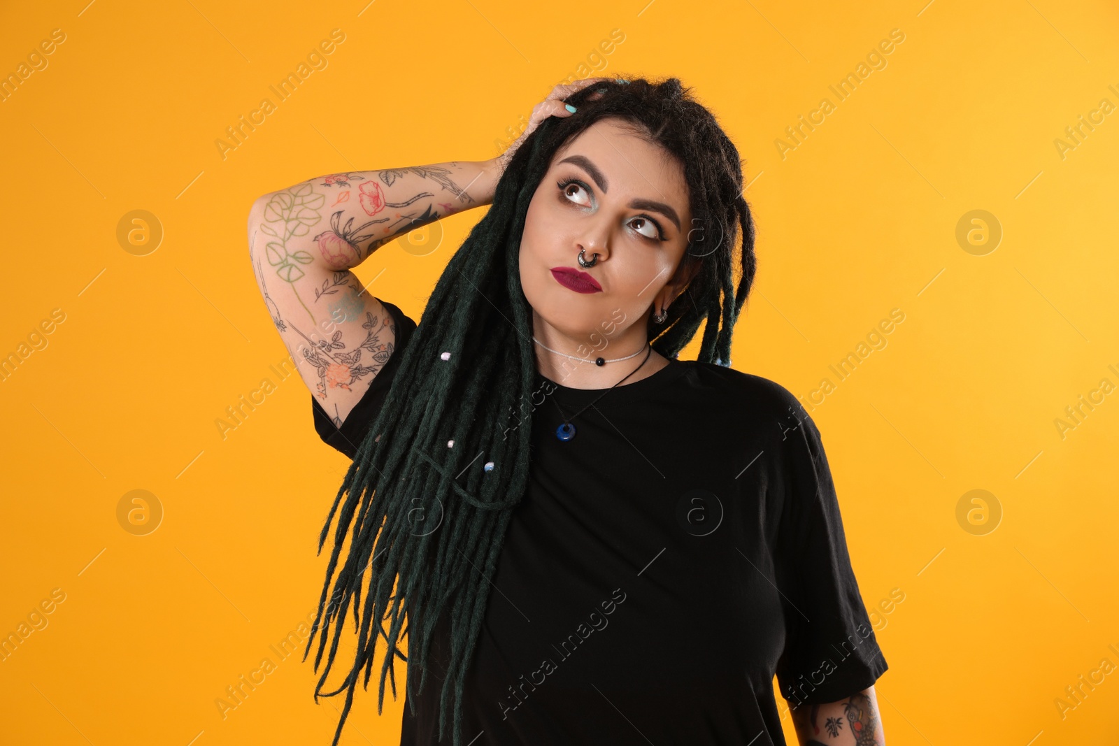 Photo of Beautiful young woman with tattoos on arms, nose piercing and dreadlocks against yellow background