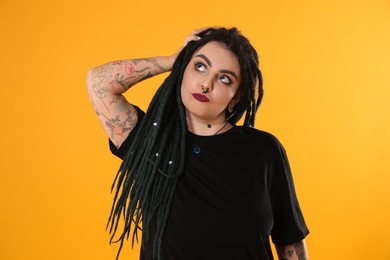 Beautiful young woman with tattoos on arms, nose piercing and dreadlocks against yellow background