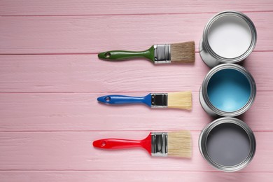 Cans of white, light blue and grey paints with brushes on pink wooden table, flat lay. Space for text