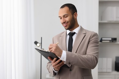 Photo of Smiling young man with clipboard writing notes in office. Lawyer, businessman, accountant or manager
