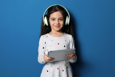 Photo of Cute little girl with headphones and tablet listening to audiobook on blue background