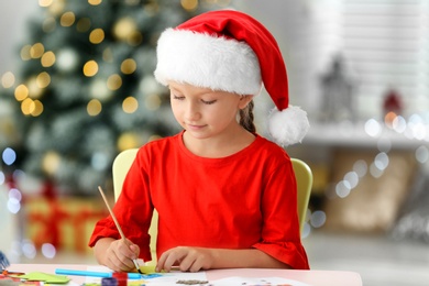 Photo of Little child in Santa hat drawing at table indoors. Christmas season