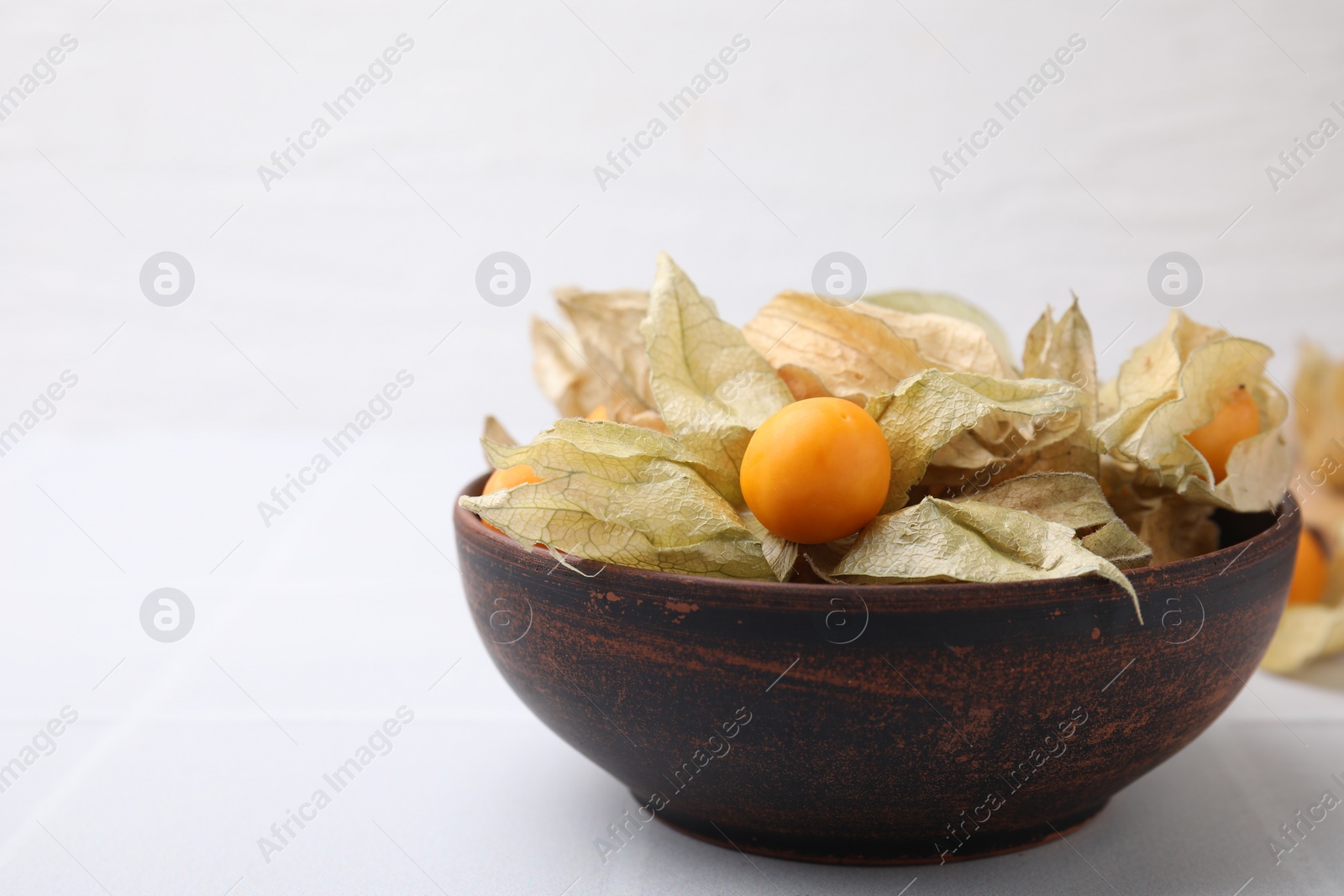 Photo of Ripe physalis fruits with calyxes in bowl on white tiled table, closeup. Space for text