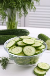 Photo of Cut cucumber in glass bowl, fresh vegetables and dill on white wooden table, closeup