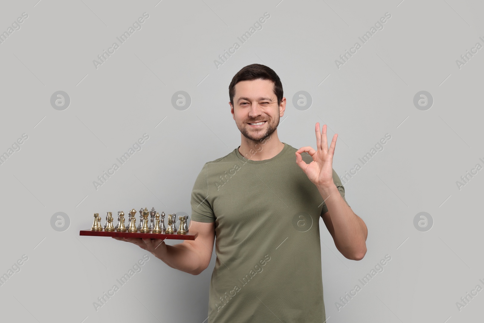 Photo of Smiling man holding chessboard with game pieces and showing OK gesture on light grey background