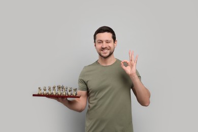 Photo of Smiling man holding chessboard with game pieces and showing OK gesture on light grey background