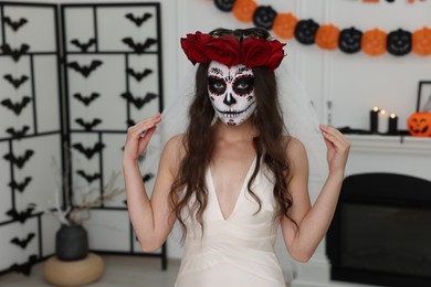 Young woman in scary bride costume with sugar skull makeup and flower crown indoors. Halloween celebration
