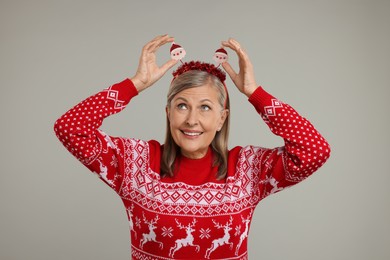 Happy senior woman in Christmas sweater and Santa headband on grey background