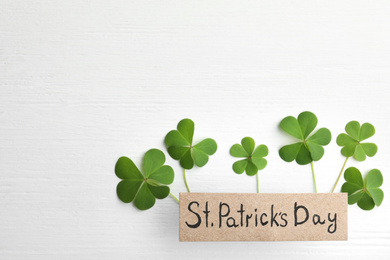 Photo of Clover leaves and card with text ST. PATRICK'S DAY on white wooden table, flat lay. Space for text