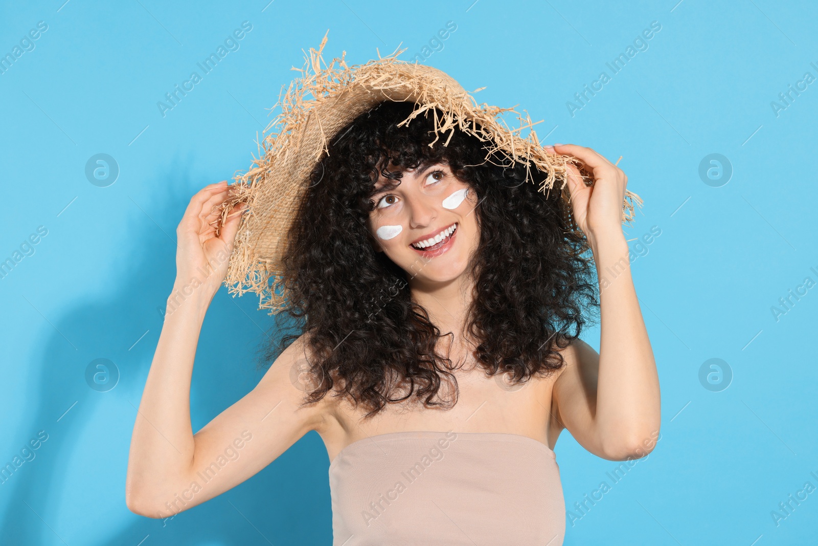 Photo of Beautiful young woman in straw hat with sun protection cream on her face against light blue background