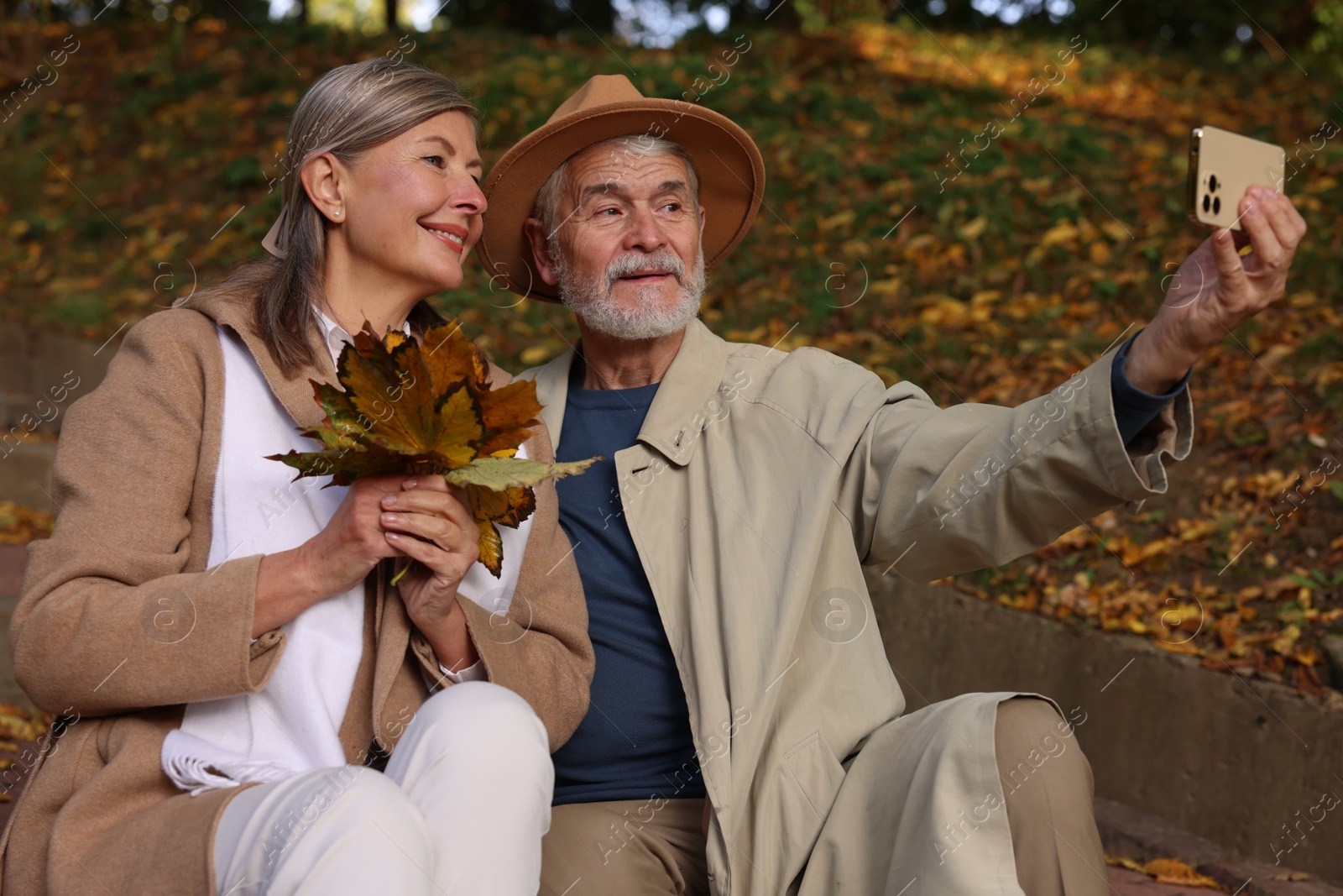 Photo of Affectionate senior couple with dry leaves taking selfie in autumn park