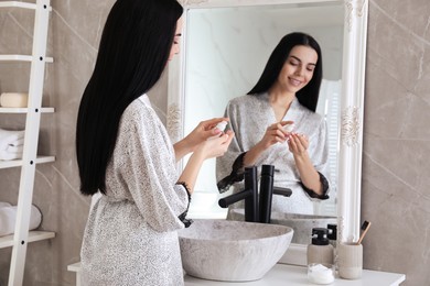 Photo of Beautiful young woman applying moisturizing cream near mirror in bathroom