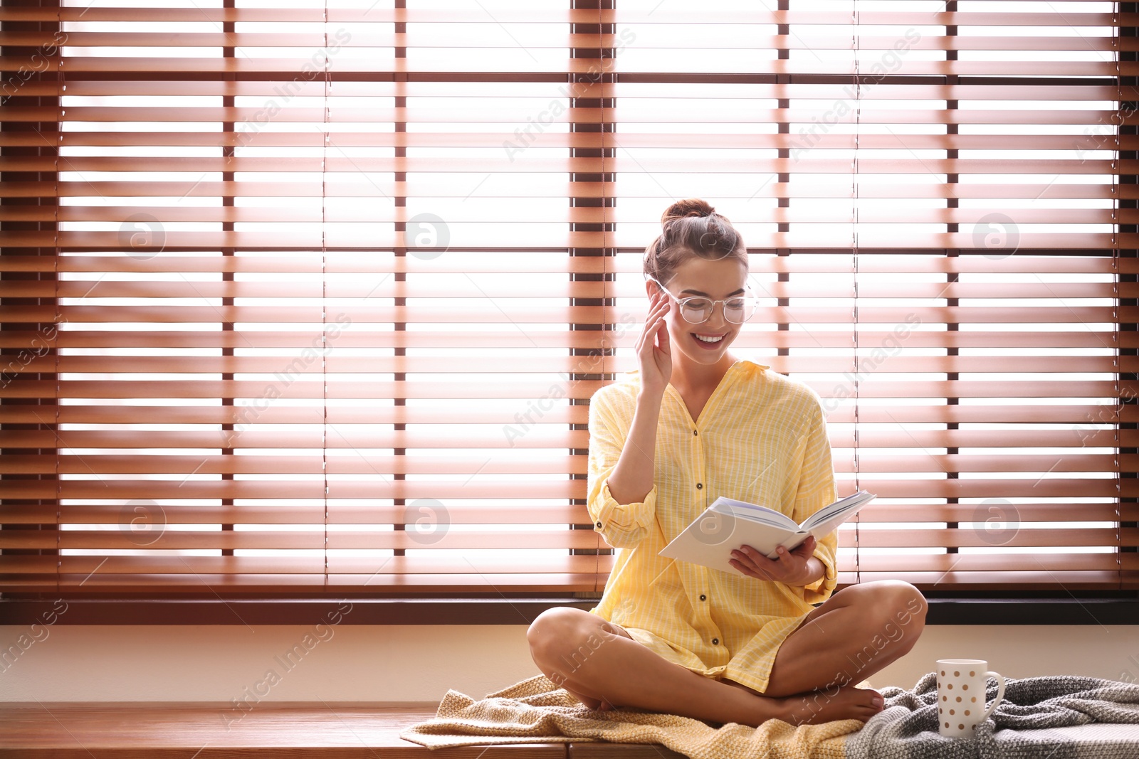 Photo of Young woman reading book near window with blinds at home. Space for text