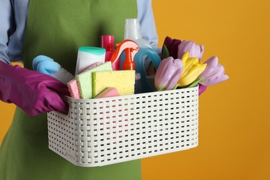 Photo of Spring cleaning. Woman holding basket with detergents, flowers and tools on orange background, closeup