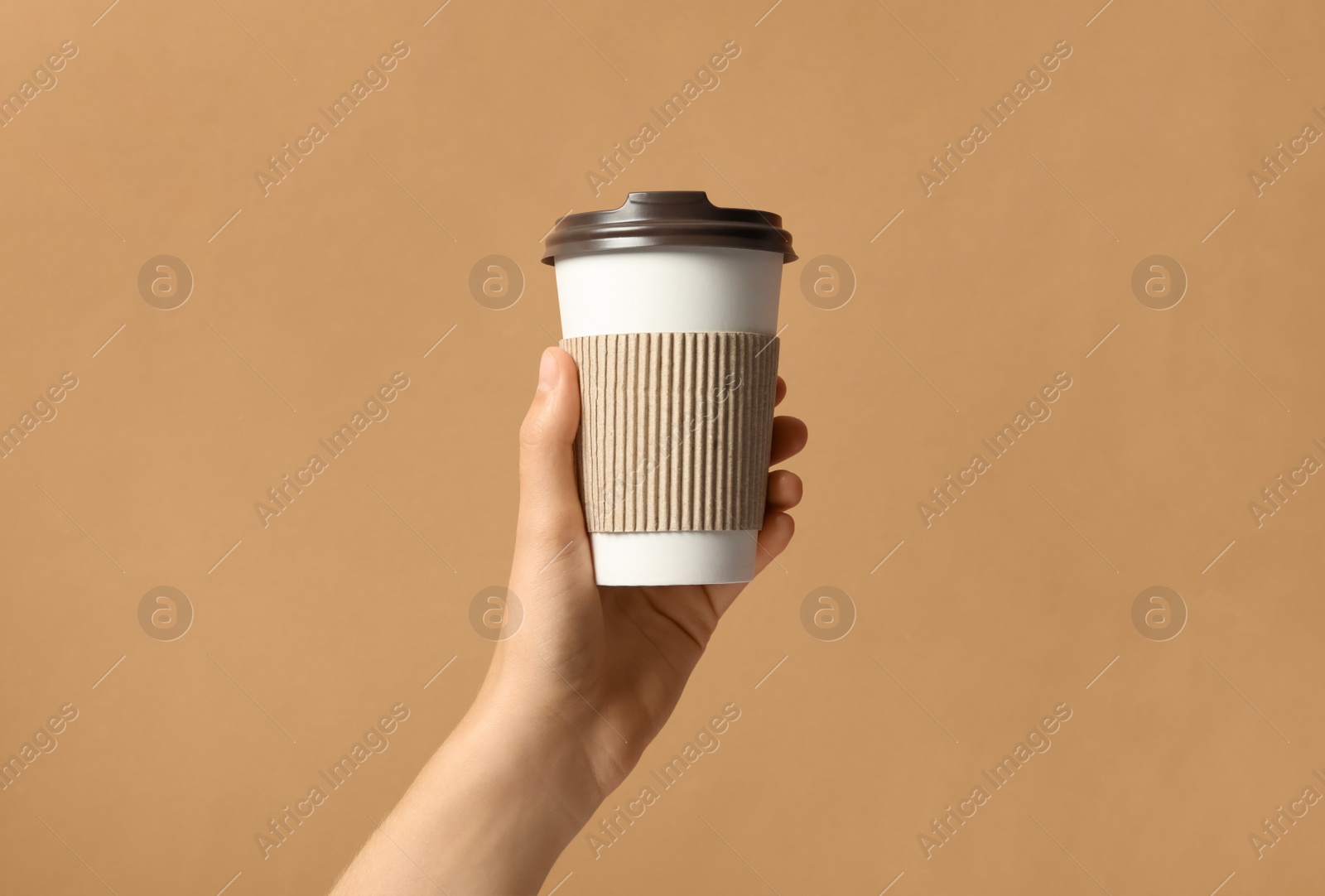 Photo of Woman holding takeaway paper coffee cup with cardboard sleeve on brown background, closeup
