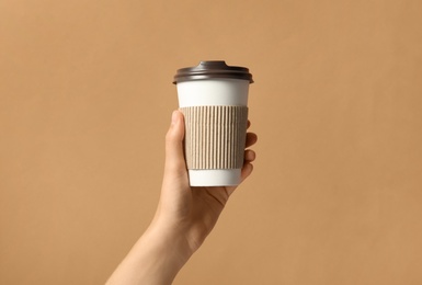 Woman holding takeaway paper coffee cup with cardboard sleeve on brown background, closeup