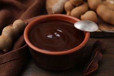 Photo of Bowl of tamarind sauce, spoon and fresh pods on wooden table, closeup