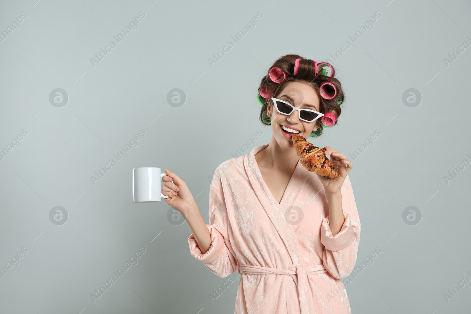 Photo of Beautiful young woman in bathrobe with hair curlers holding croissant and cup of drink on light grey background